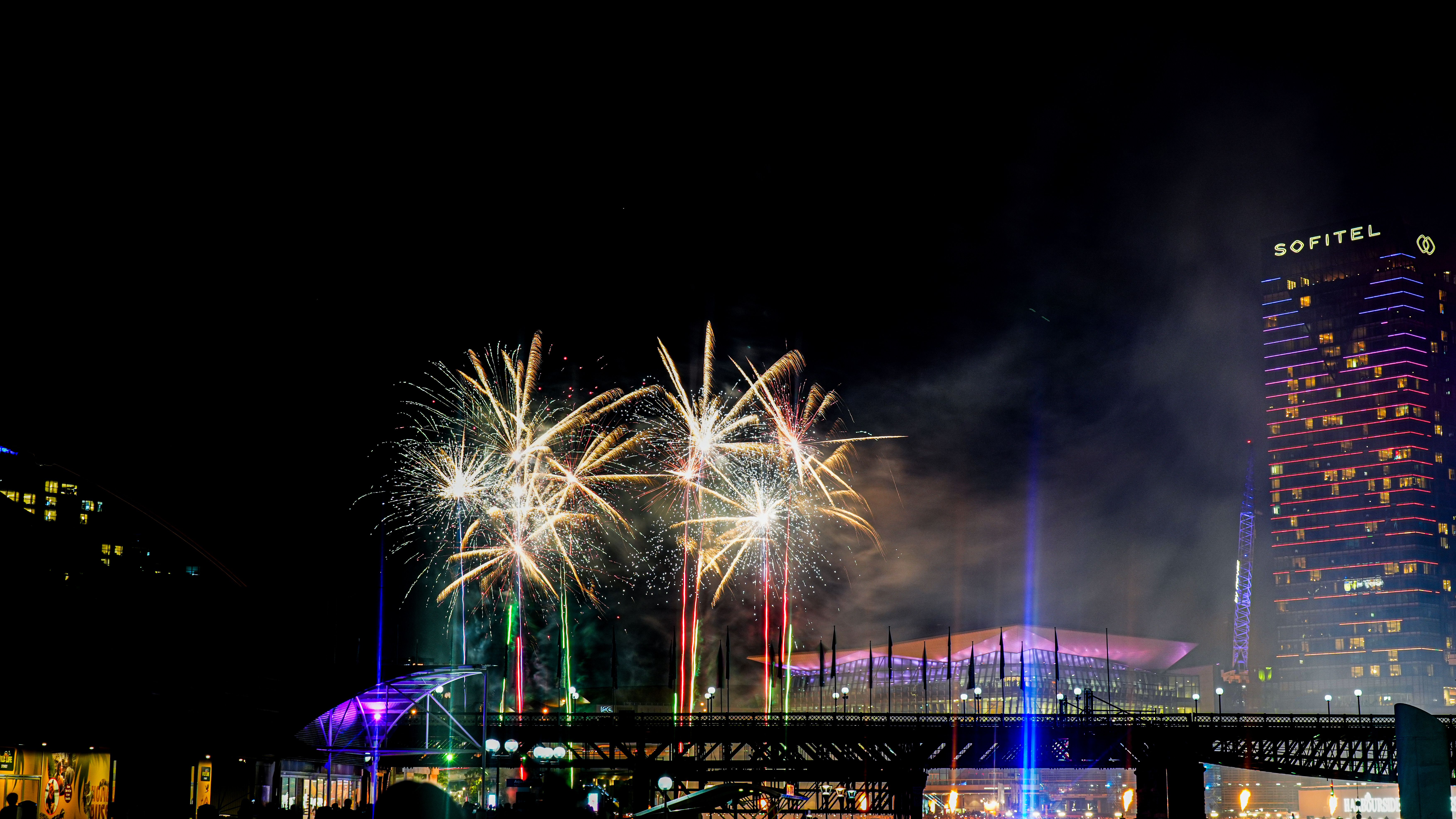 Fireworks bursting over a lit bridge at night, with a tall hotel tower marked Sofitel on the right
