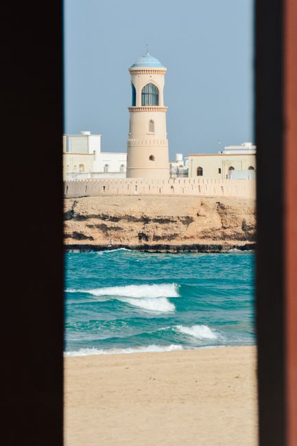 Scenic view of a lighthouse through a window with beach and ocean in Muscat, Oman.