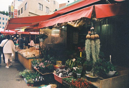 Lively outdoor flower market with various plants and bustling shoppers under red awnings.