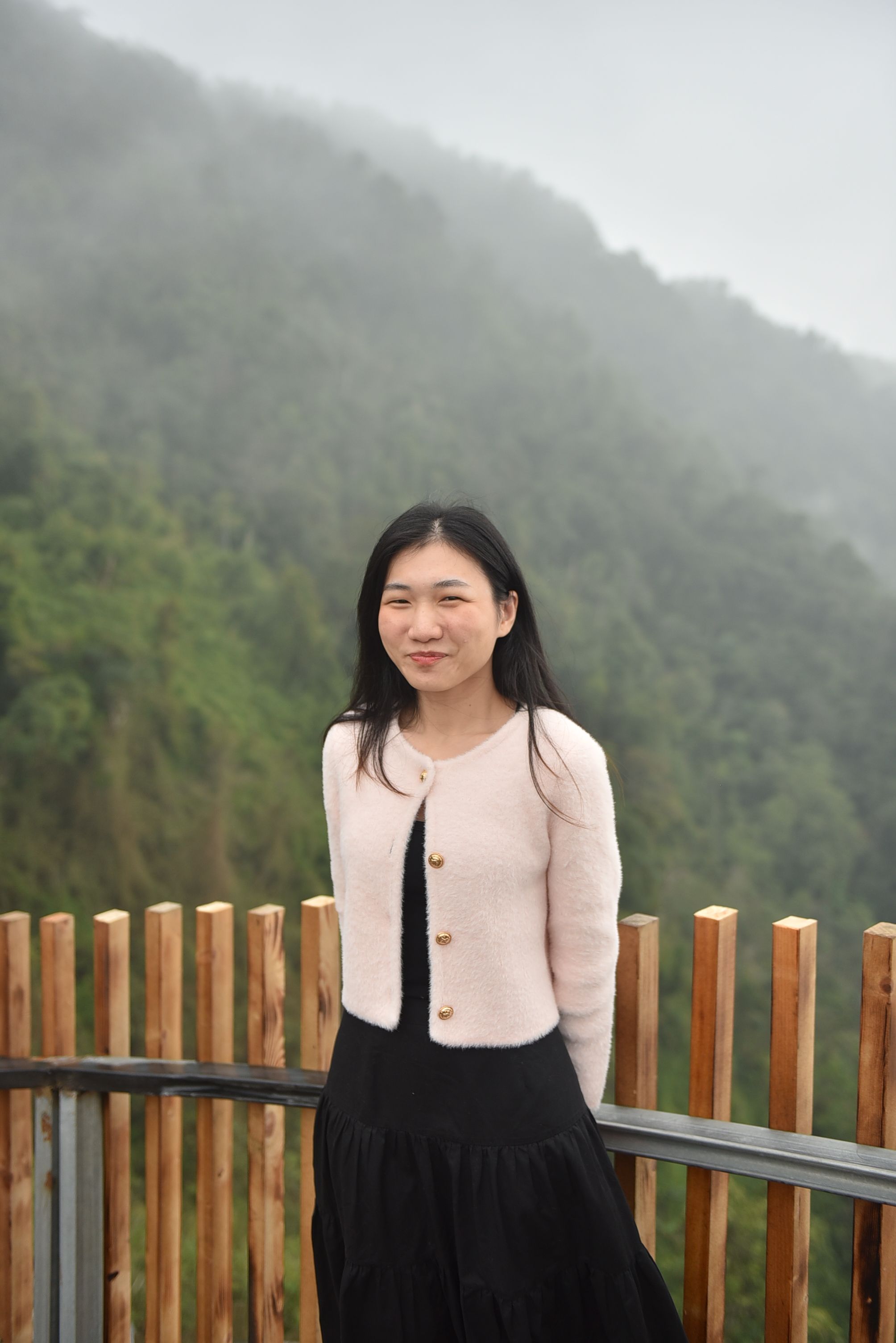 A young woman smiles against a misty forest backdrop, wearing casual attire. Fog adds a serene atmosphere.