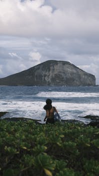 A woman stands by the ocean with a view of a large mountain under cloudy skies.
