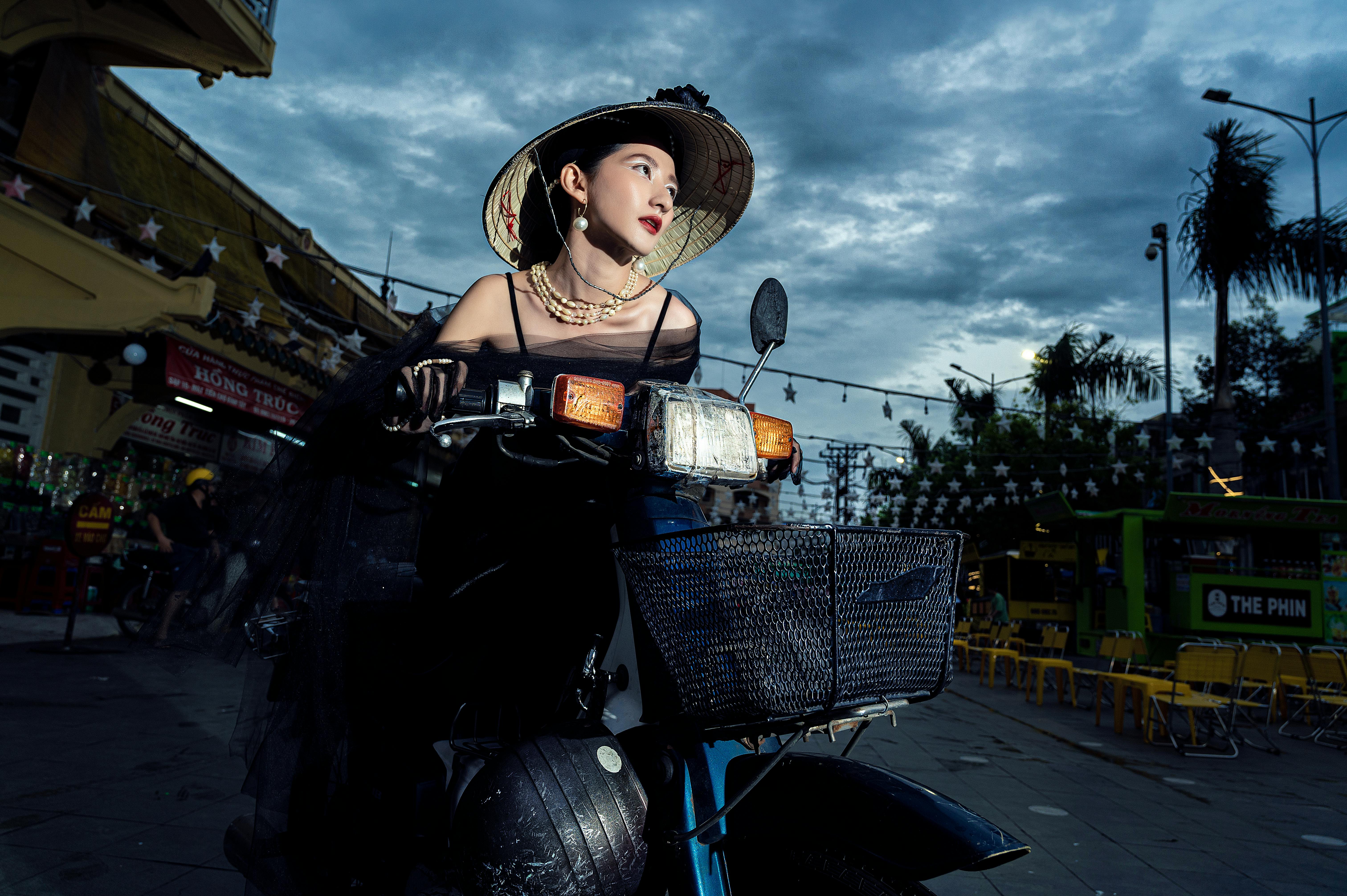 Free Elegant woman on a vintage scooter in an urban setting at dusk, wearing a hat and pearl necklace. Stock Photo