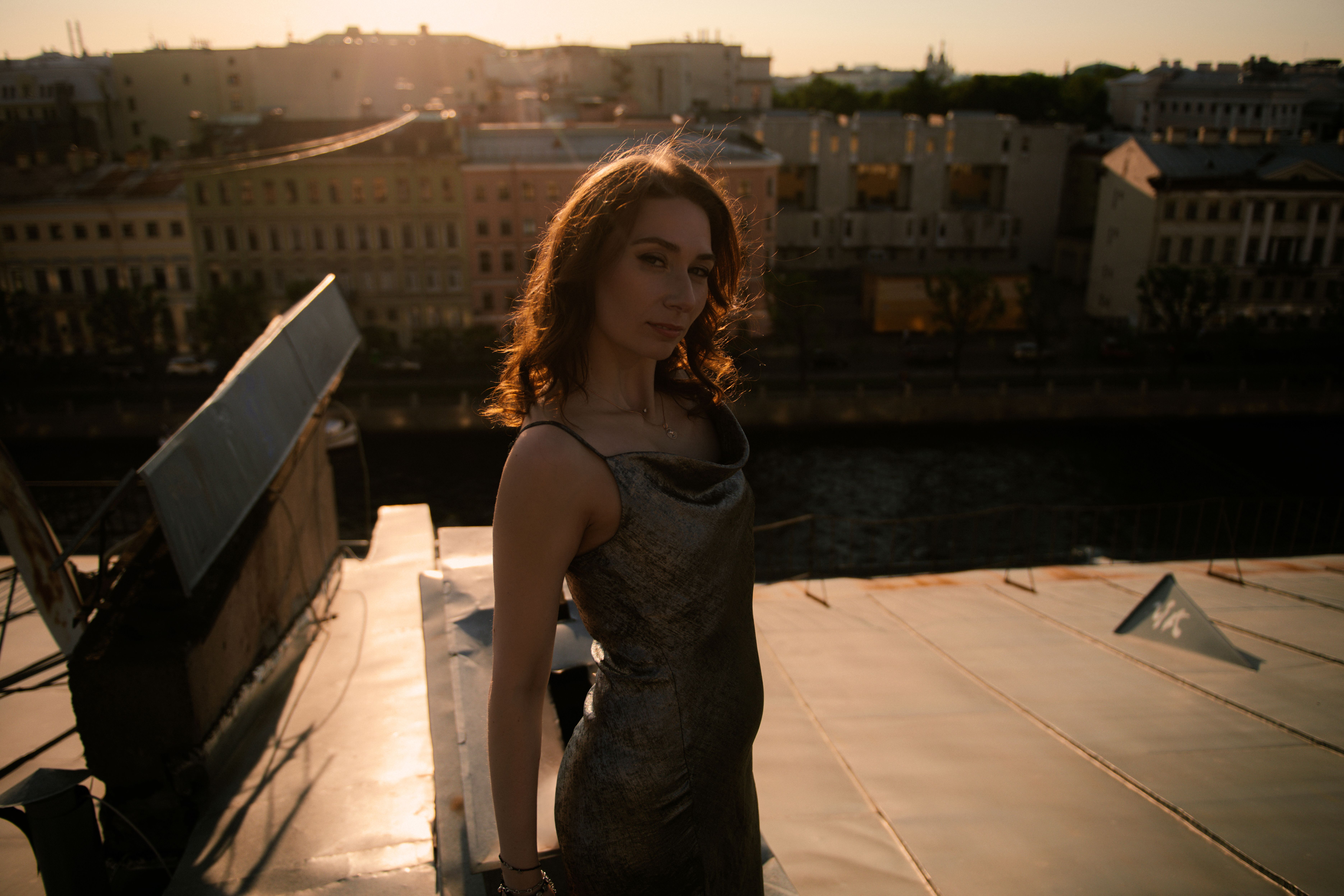 A graceful woman standing on a rooftop with a cityscape view at sunset.