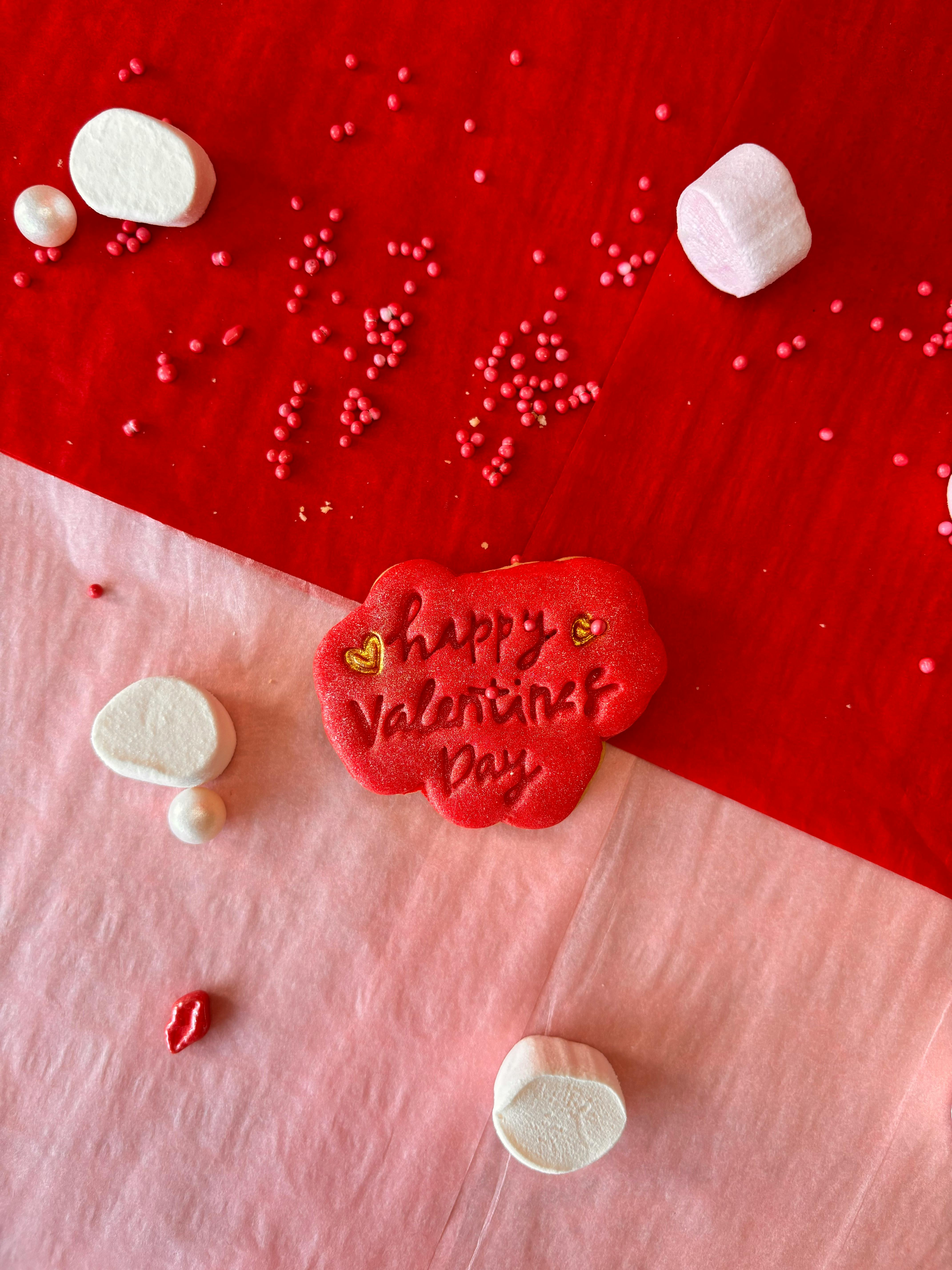 Red Valentine's Day cookie surrounded by white marshmallows and red decorations on a festive background.