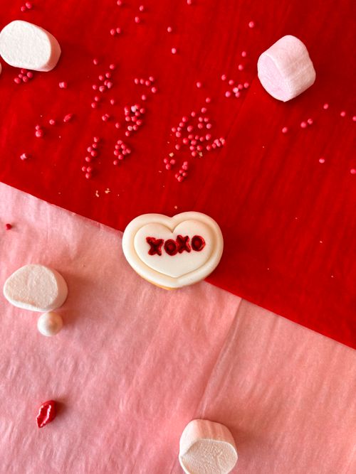 Free Heart-shaped cookie with 'XOXO' text, surrounded by marshmallows and sprinkles on red and pink fabric. Stock Photo