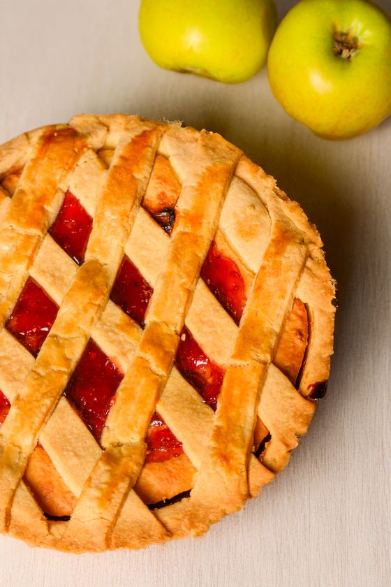 Golden lattice-top pie with glossy red strawberry apple filling beside two green apples