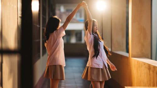 Free Two Vietnamese schoolgirls dance joyfully in a sunlit corridor, capturing a moment of youth and happiness. Stock Photo