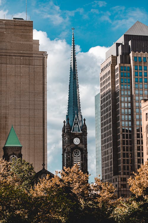 Old tower with spire and clocks in Gothic style located among modern high rise buildings on city street against cloudy blue sky