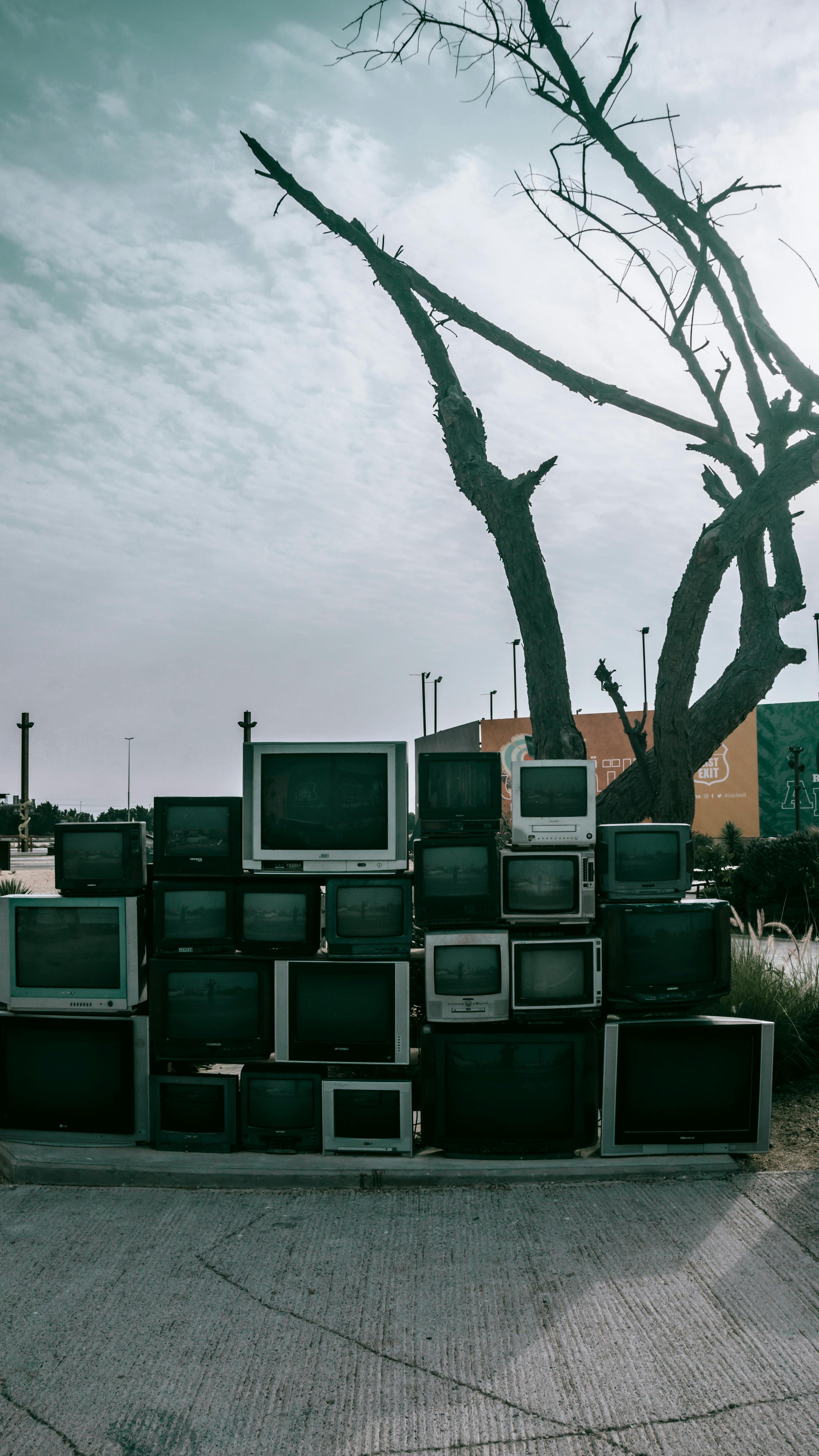A collection of vintage televisions stacked outdoors under a barren tree, capturing a juxtaposition of technology and nature.