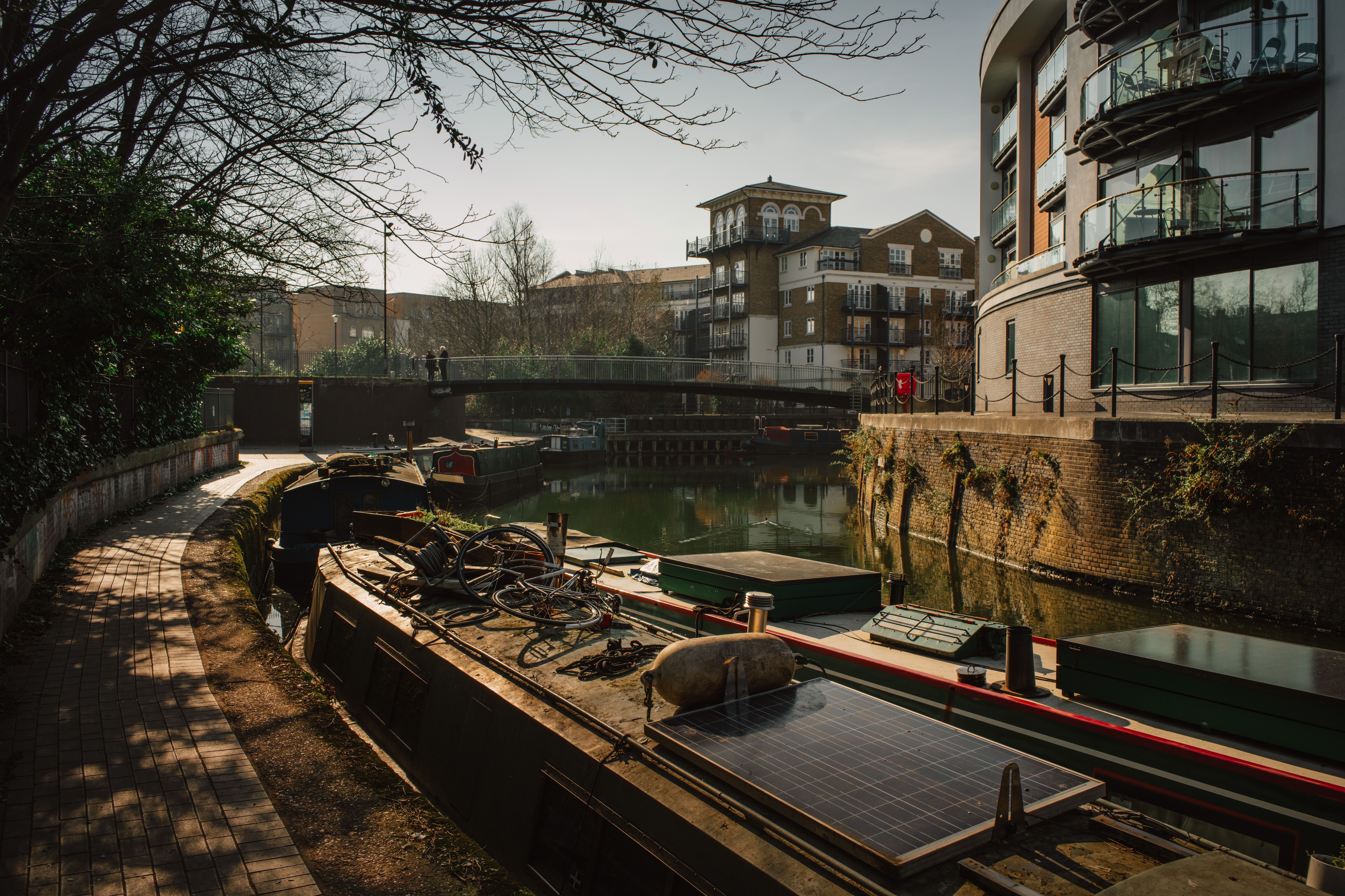 Sunlit view of canal with docked narrowboats and modern buildings in urban setting.