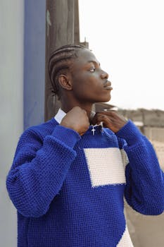 A young man in Lagos, Nigeria, wearing a blue sweater adjusts his necklace in a confident pose.