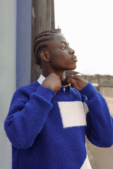 A young man in Lagos, Nigeria, wearing a blue sweater adjusts his necklace in a confident pose.