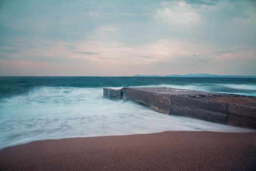 Tranquil scene of waves crashing on a pier at a Greek beach, captured in long exposure during sunset.