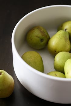 A collection of fresh green pears placed in a minimalist white bowl on a dark surface.