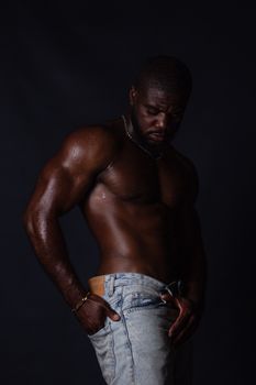 A muscular man with sweat on his body poses dramatically in jeans against a dark background.