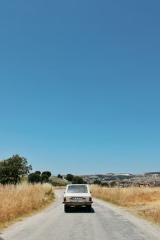 A vintage car travels down a rural road surrounded by fields under a clear blue sky.