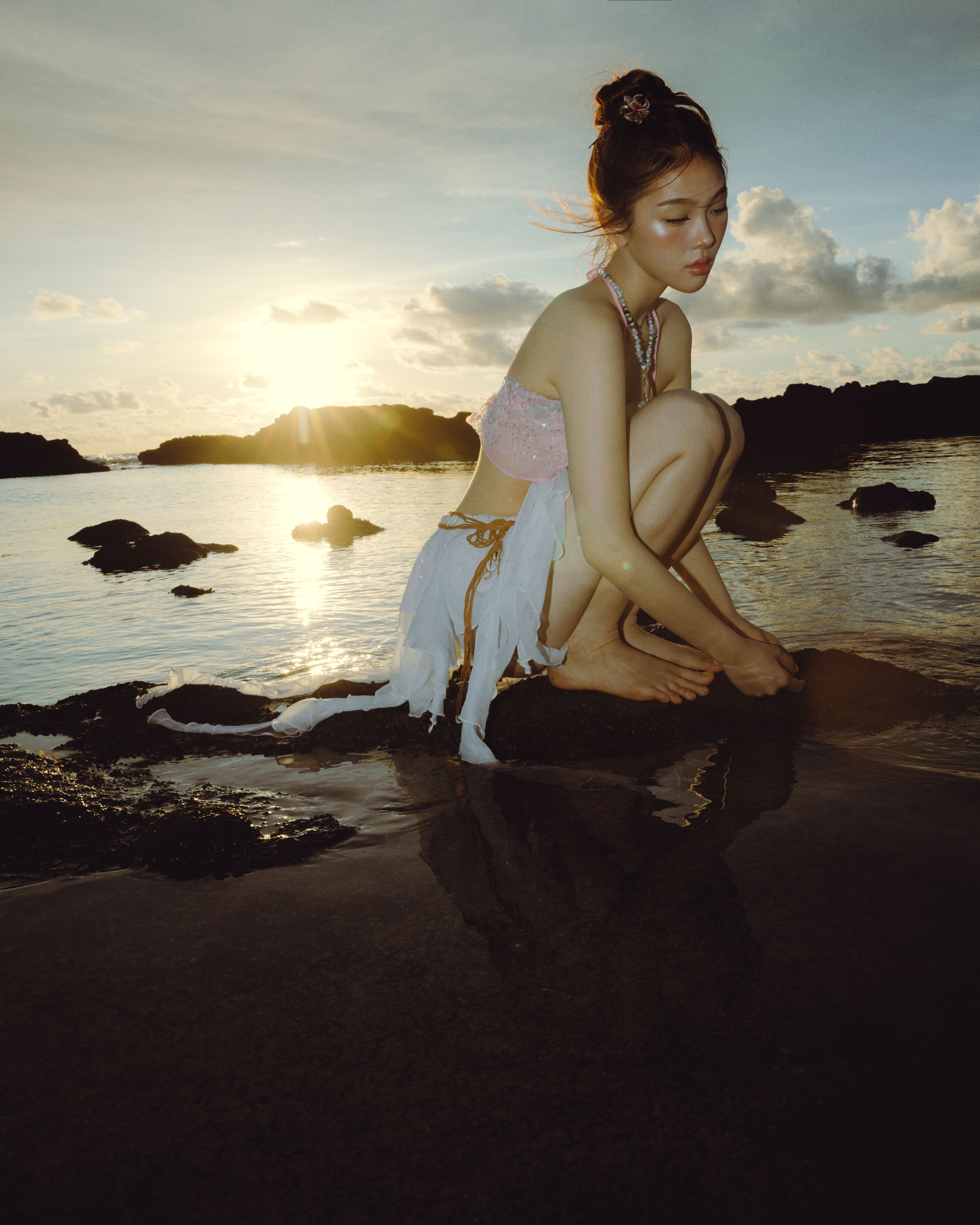 A young woman in a unique outfit crouches by a rocky beach at sunset, reflecting in water.
