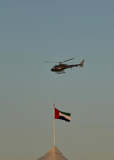A military helicopter flies above the UAE flagpole against a clear sky.