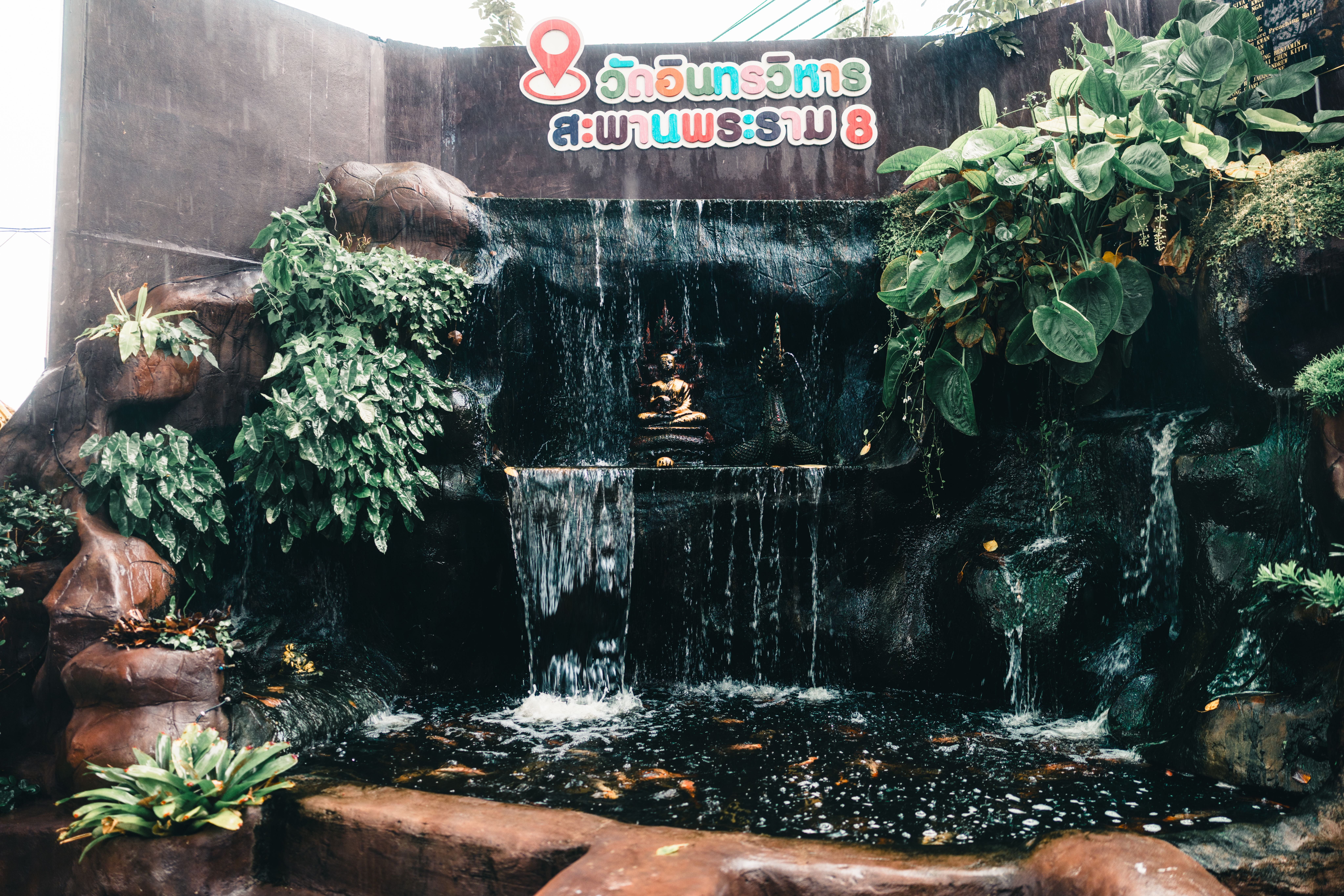 Tranquil scene of a waterfall with Buddha statue surrounded by lush greenery in Bangkok, Thailand.