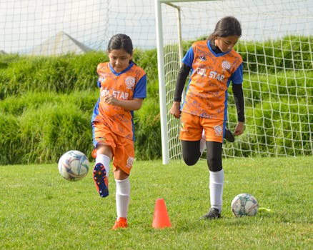 Two young girls in soccer uniforms practicing on a grassy field with a goalpost.