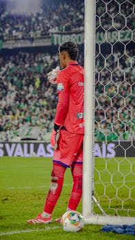 Soccer goalkeeper in pink kit drinking water during a lively match in a stadium.