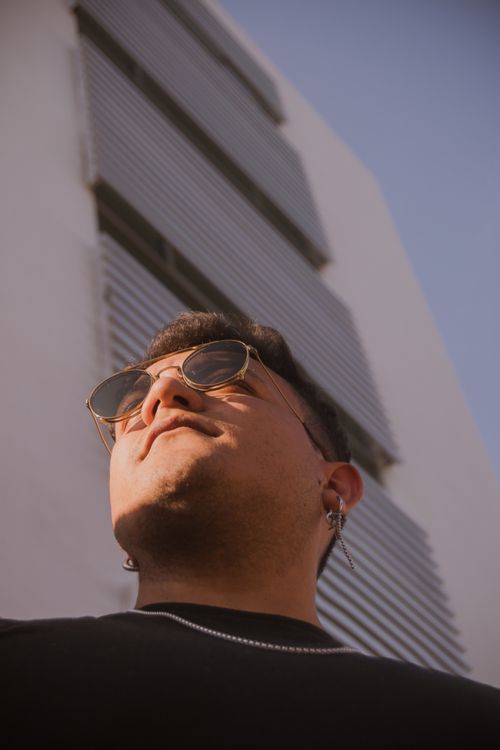 Free Young man wearing sunglasses looks up confidently against modern building backdrop. Stock Photo
