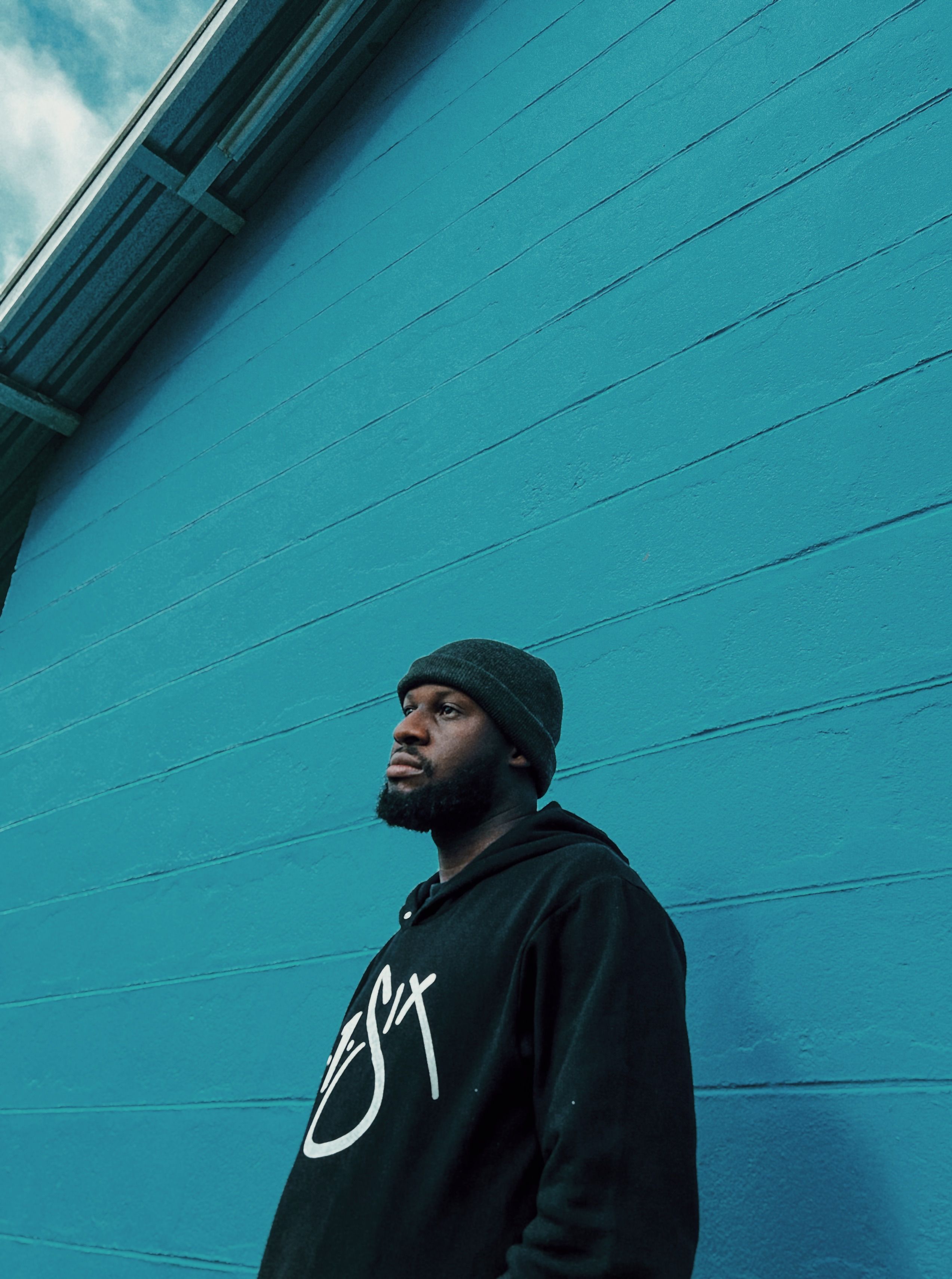 Young man in black hoodie and beanie standing against a bright blue wall, looking thoughtful.