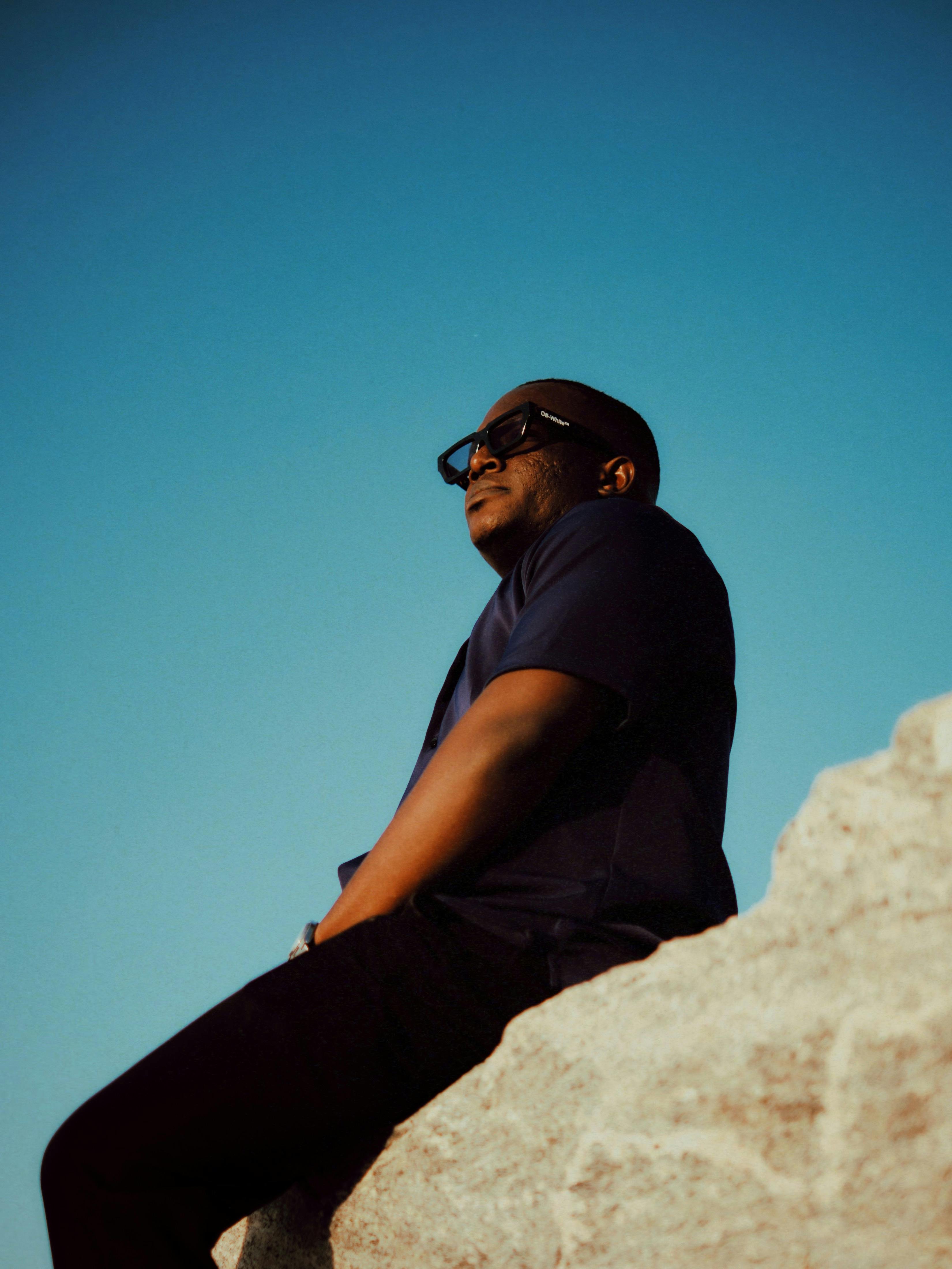 A man in sunglasses enjoying a serene moment on a rock during sunset, against a clear sky.