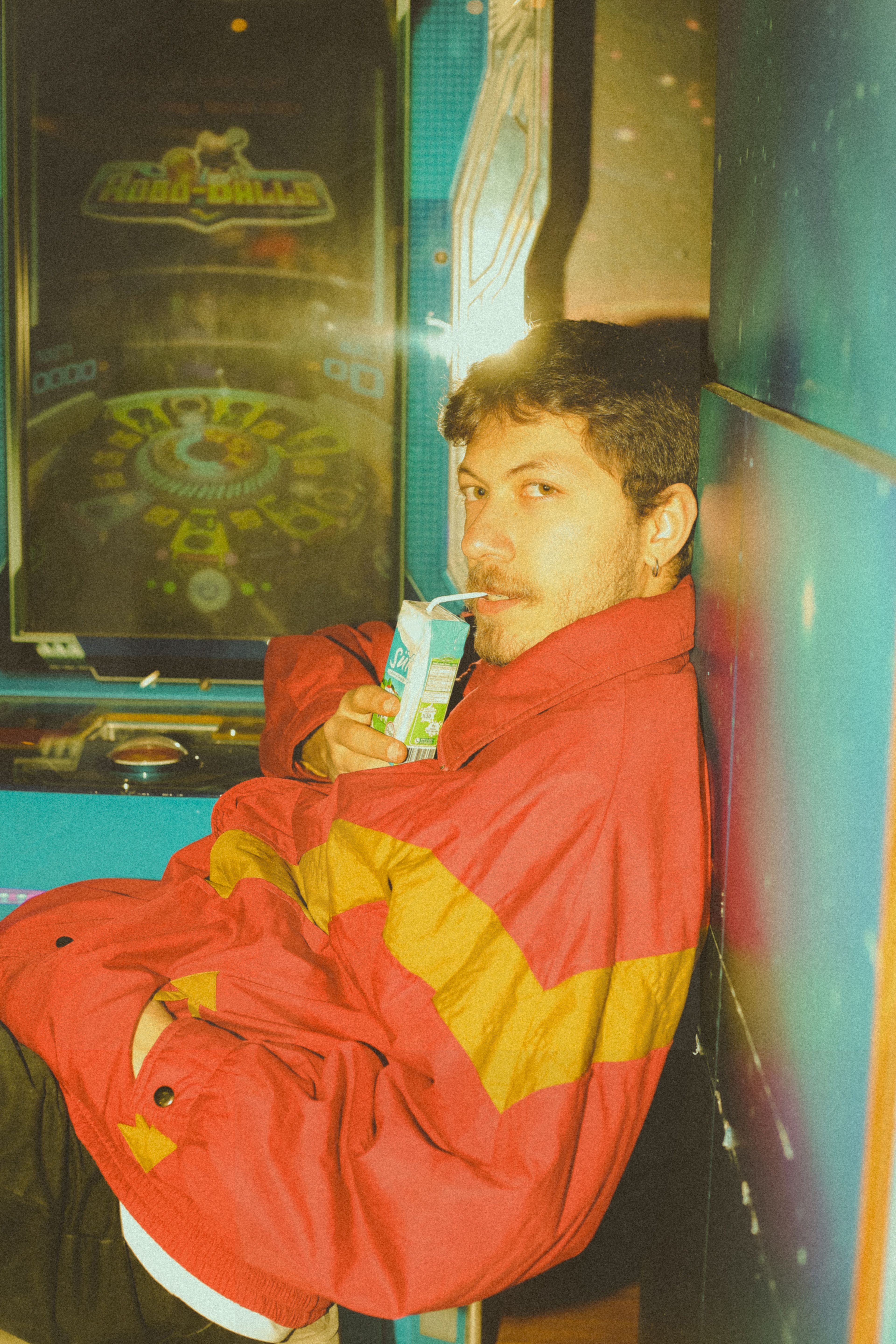 Man in vintage jacket drinks juice while enjoying retro arcade vibes.