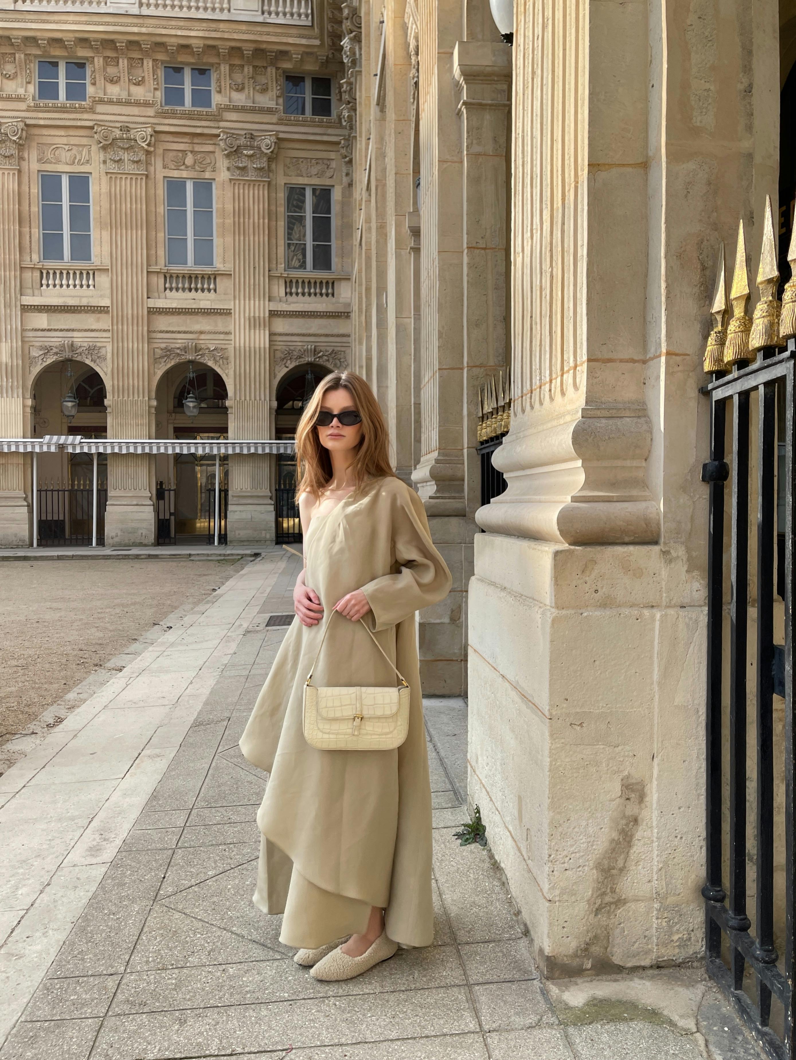 Chic woman in beige dress and sunglasses poses by a historic building, radiating timeless fashion.