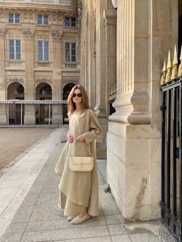 Chic woman in beige dress and sunglasses poses by a historic building, radiating timeless fashion.
