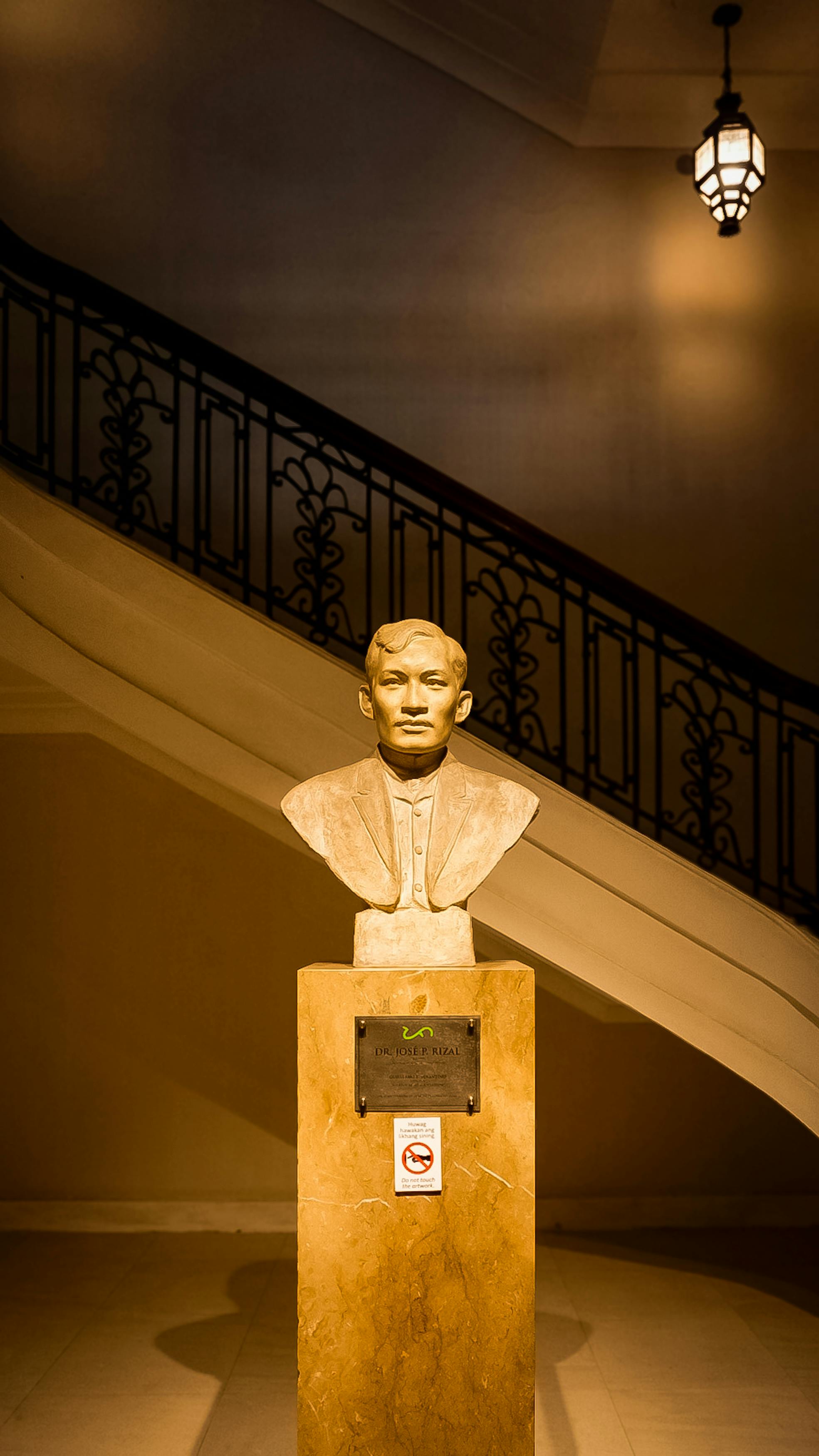 grátis Busto de uma figura histórica proeminente, sob iluminação quente de museu com uma escadaria intrincada ao fundo. Foto profissional