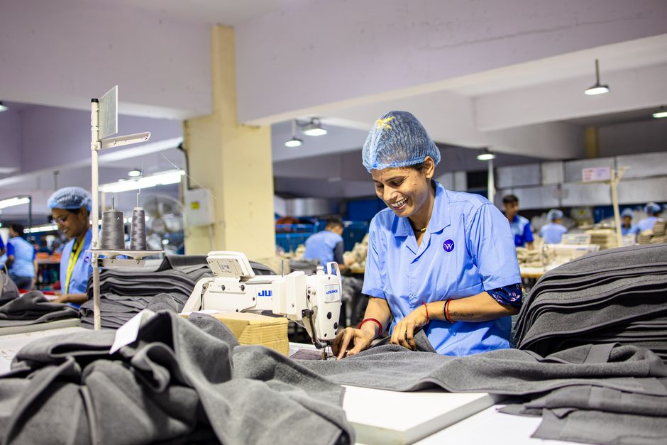Female worker operates sewing machine in bustling textile factory, smiling and focused.