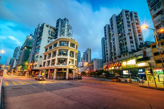 A vibrant street scene featuring a historic building in Kowloon, Hong Kong, during evening twilight.