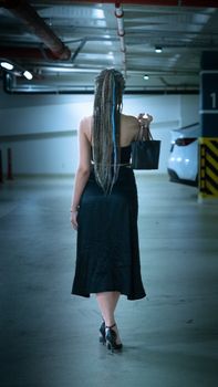 Fashionable woman with dreadlocks in a black dress walking in an İzmir parking garage.