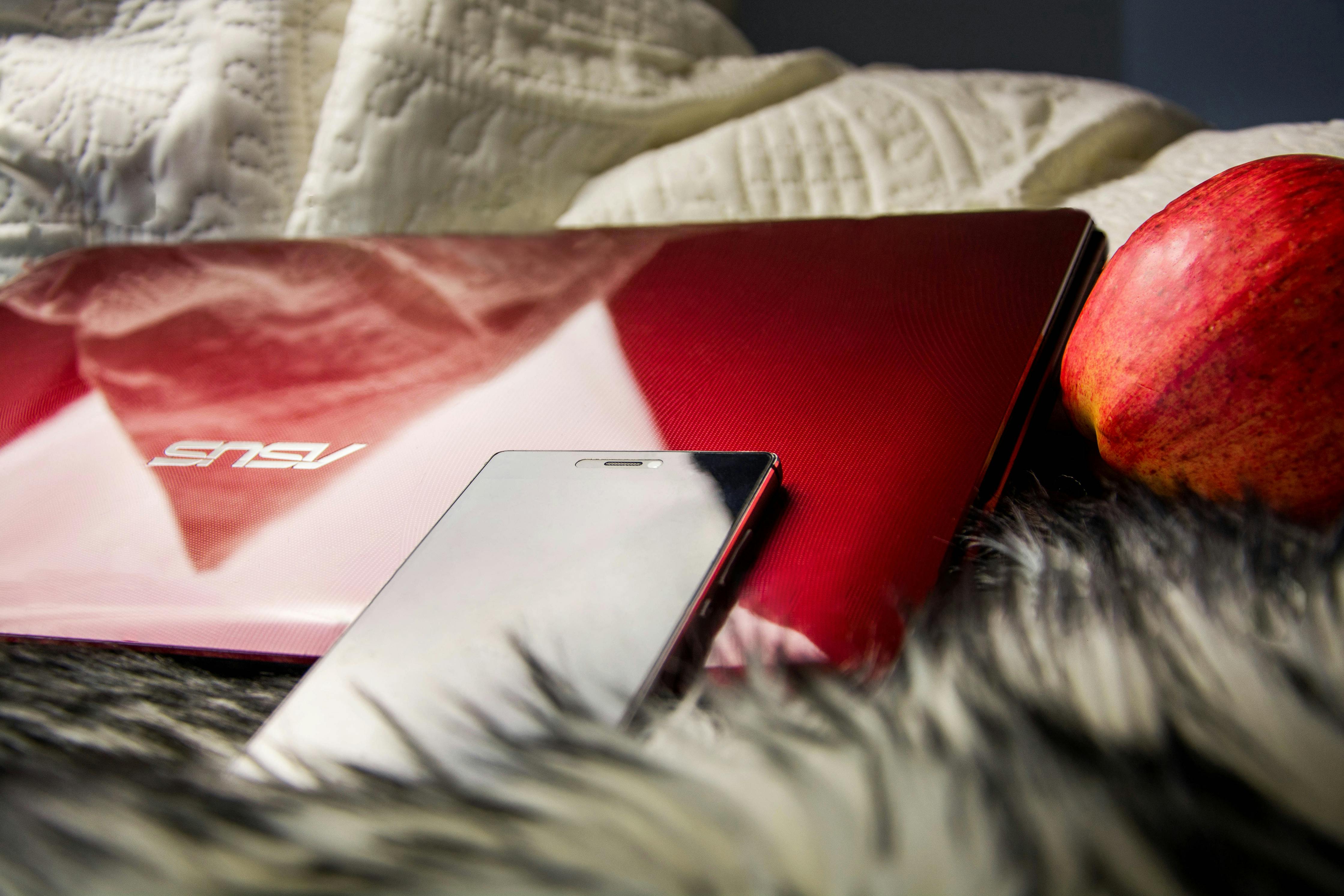 Red laptop with smartphone and apple on cozy blanket, creating a relaxed workspace vibe.