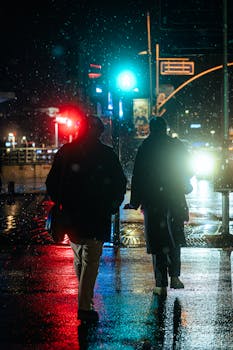 Two silhouetted figures walk through a rainy city at night, illuminated by streetlights and reflections.