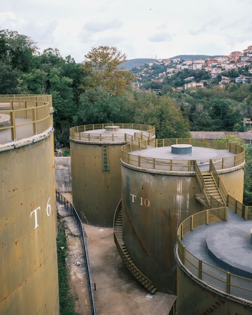 Free Aerial view of large, rust-colored storage tanks surrounded by greenery and a distant townscape. Stock Photo