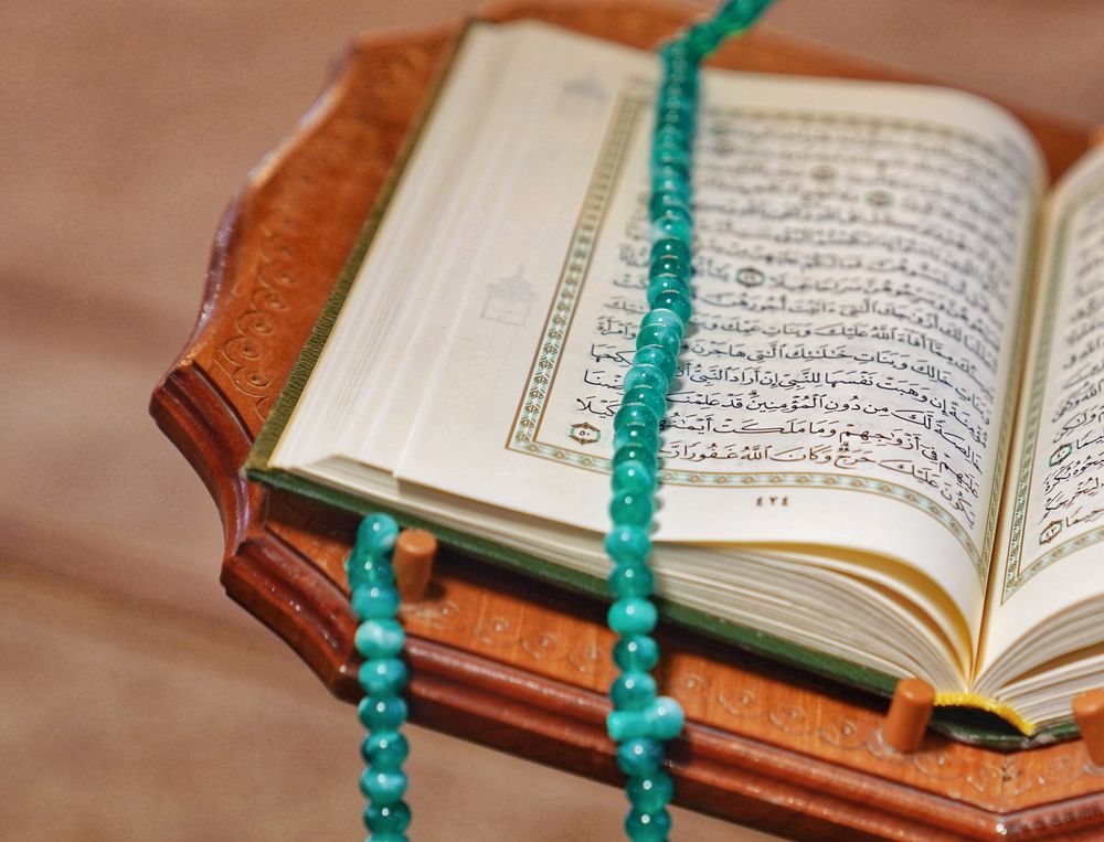 An open Qur'an resting on a wooden stand with prayer beads, softly lit