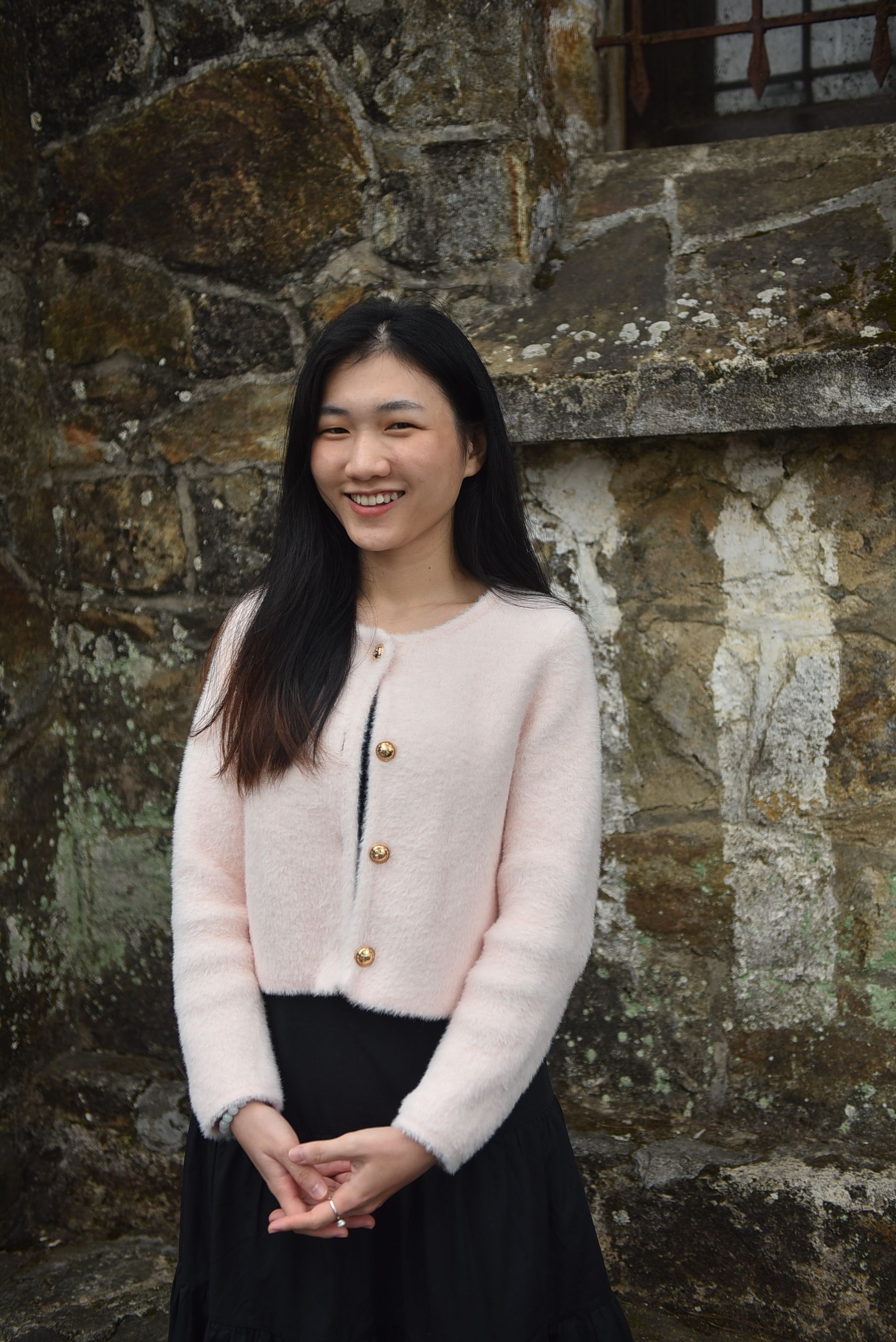 Young woman in a pink sweater smiling against a rustic stone wall.