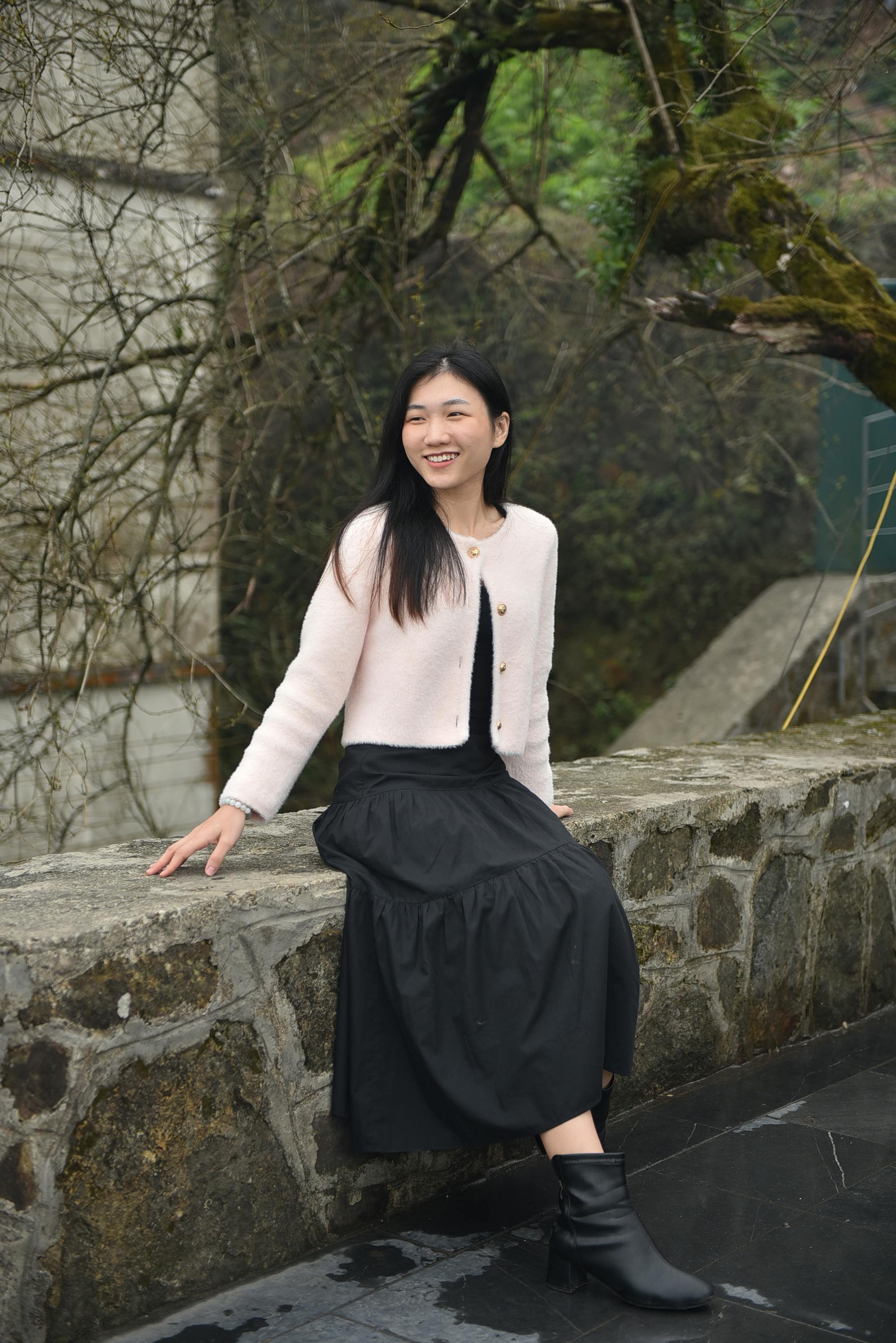 A young woman in stylish clothing sits on a stone wall outdoors, radiating joy.