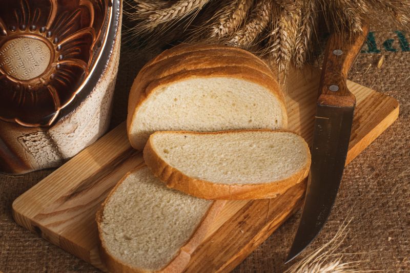 Sliced rustic bread on a wooden board with wheat and a decorative pot.