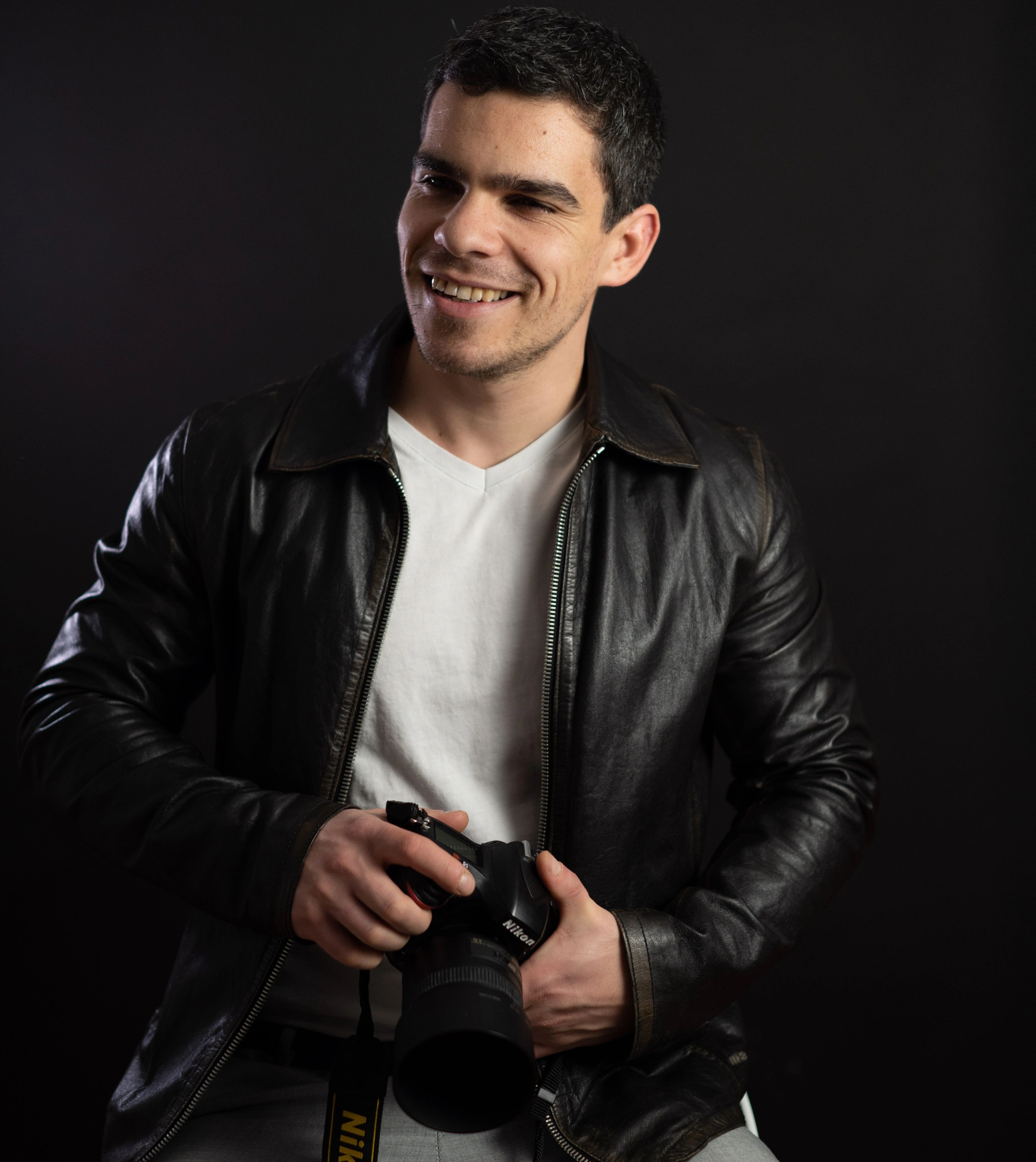 Smiling male photographer in leather jacket holding camera in a studio setting.