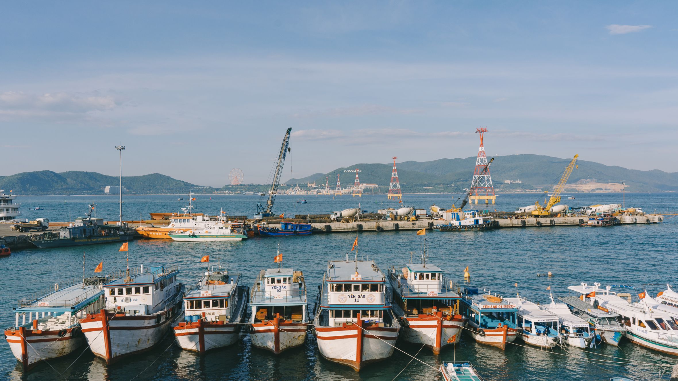 A vibrant harbor scene with fishing boats anchored and cranes in the background under a clear sky.