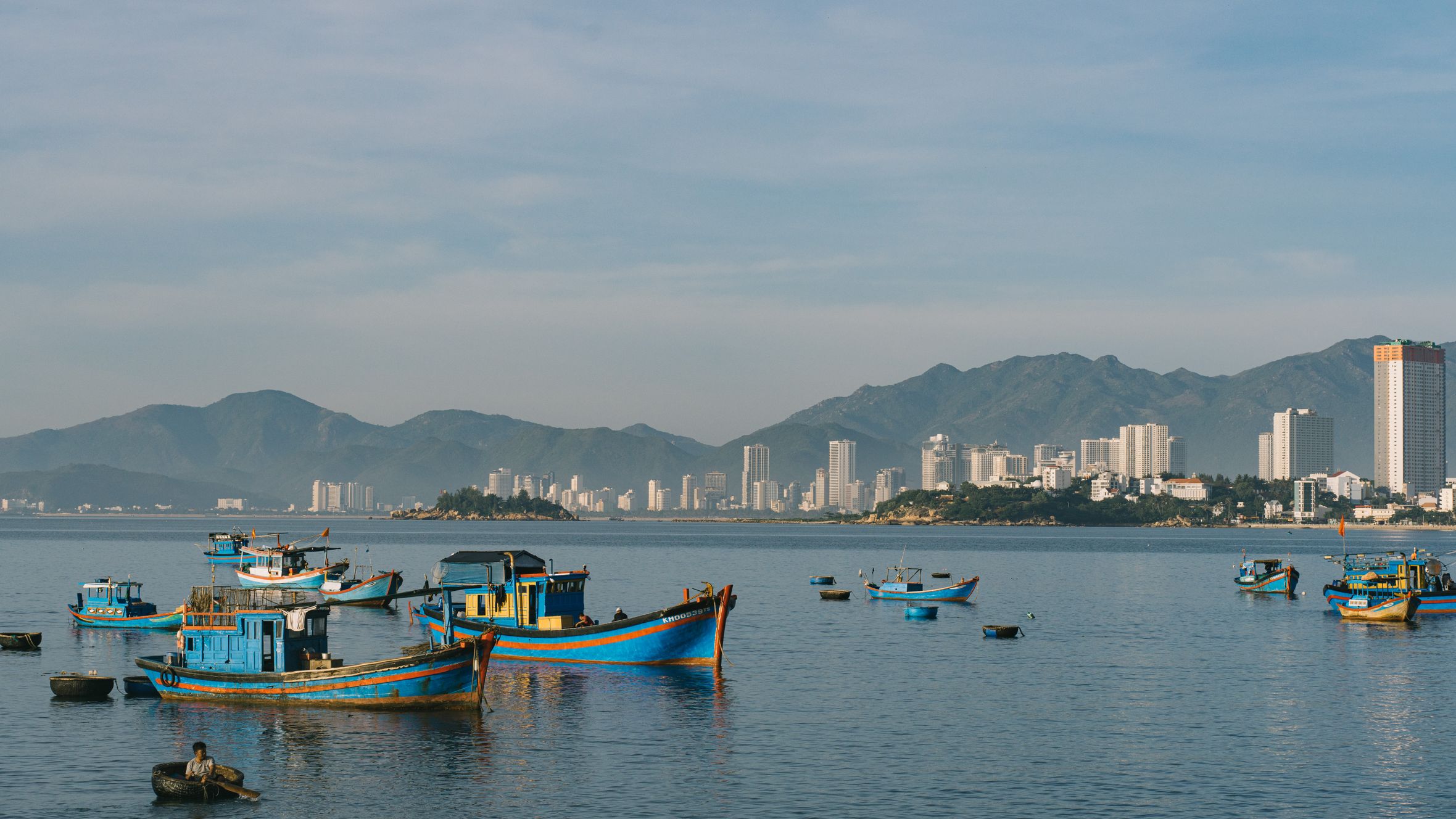 Vibrant fishing boats anchored with a city skyline and mountains in the background.