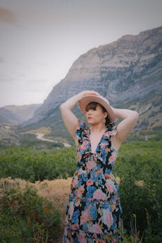 A woman poses in a floral dress and hat against a picturesque mountain backdrop, showcasing a blend of fashion and nature.