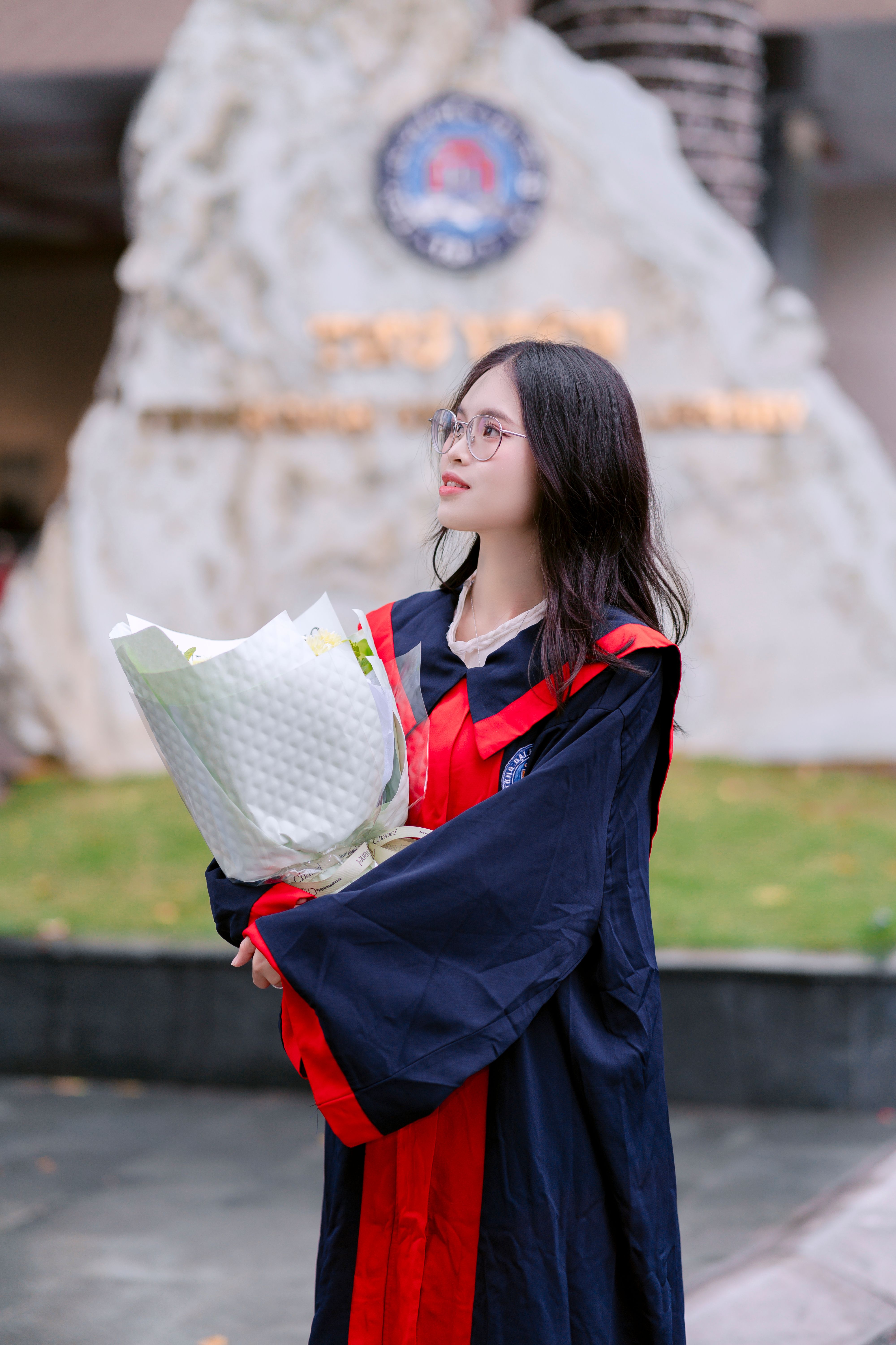 A graduate in a cap and gown celebrates with a bouquet, outdoors at a recognizable university landmark.