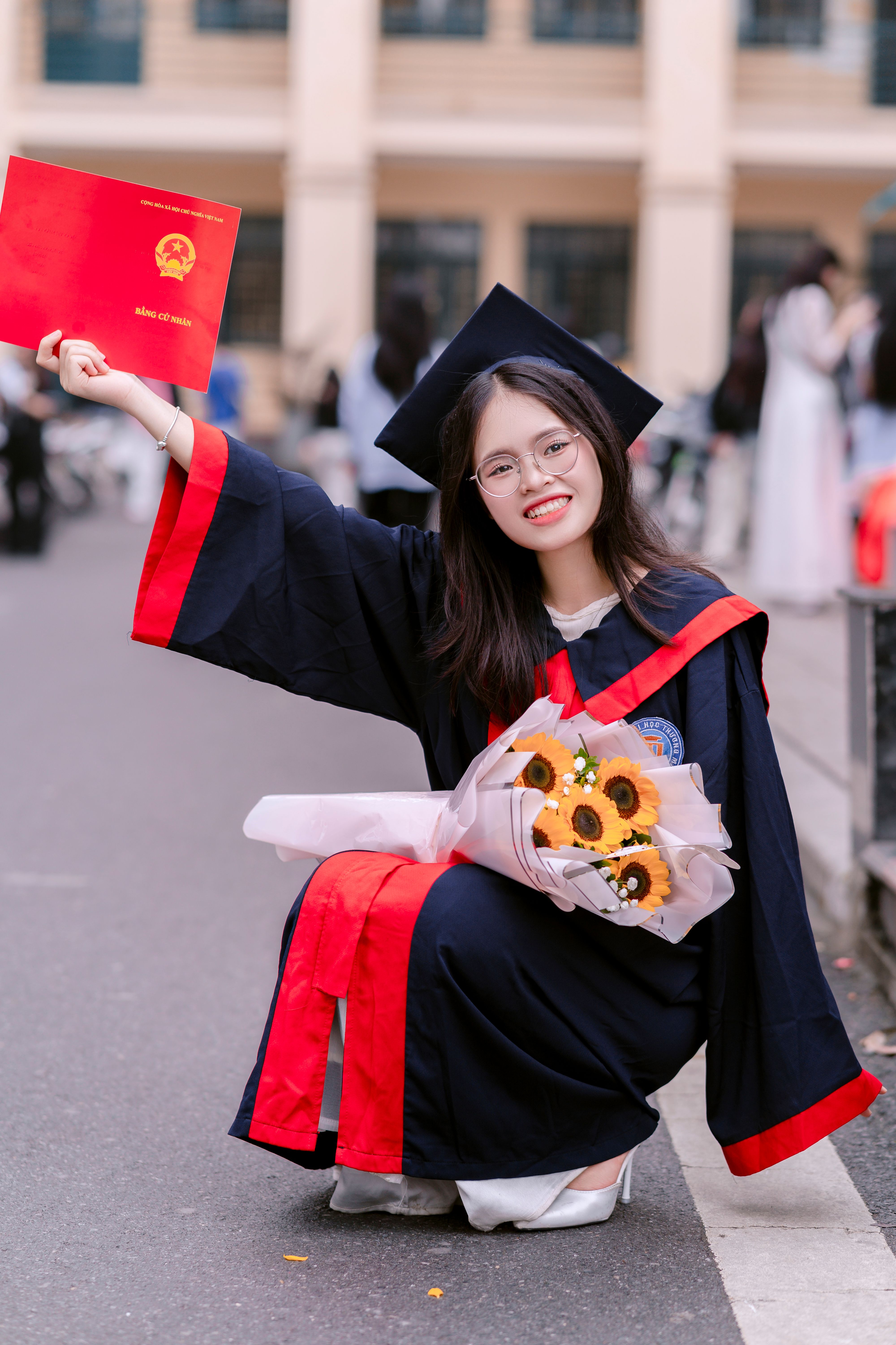 Young graduate in cap and gown celebrating with diploma and flowers outdoors.