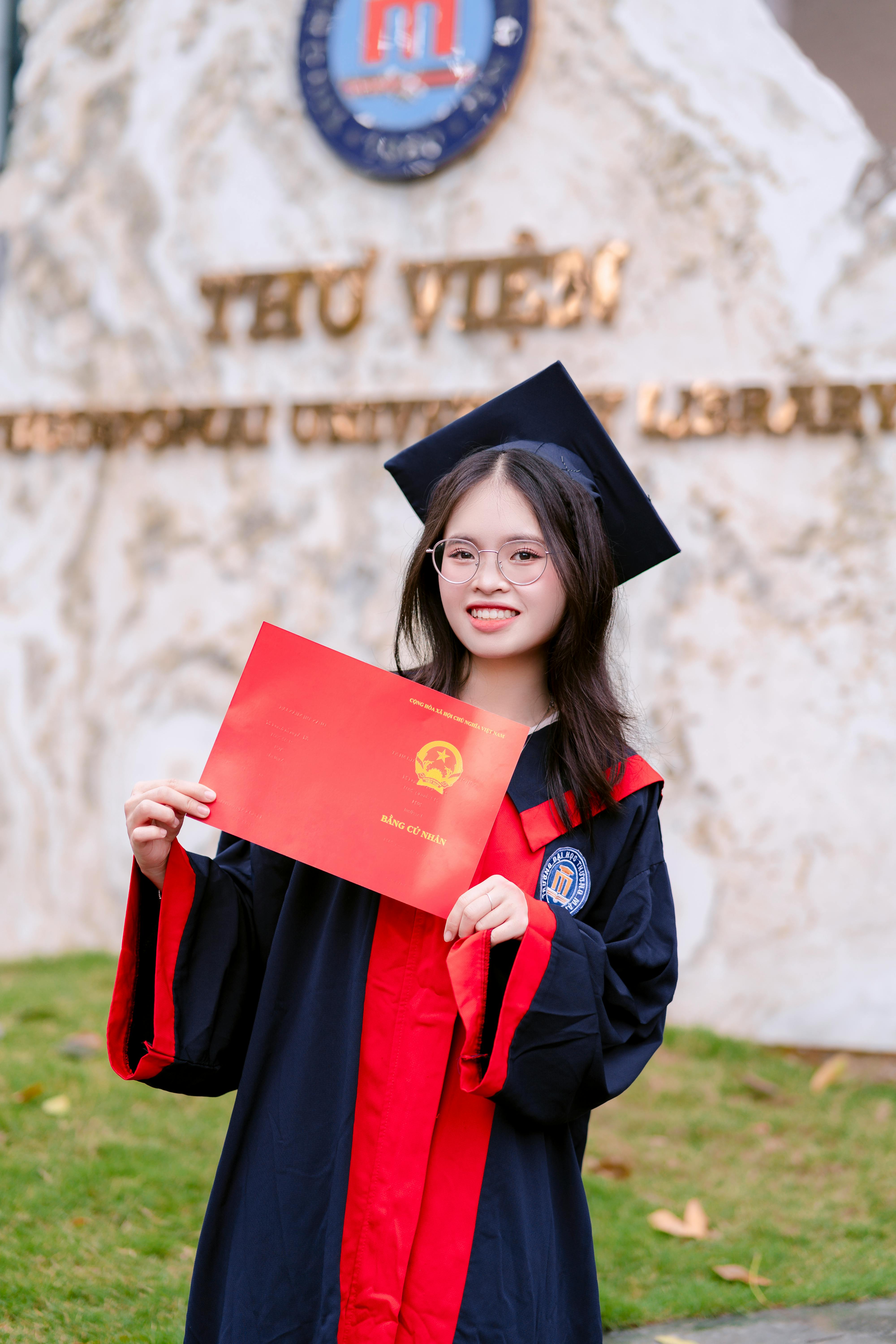 Young woman in graduation gown holding diploma outdoors with university background.