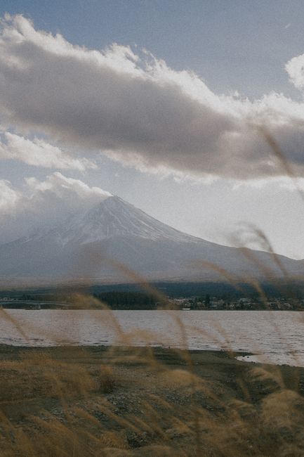 Mount Fuji towering amidst a cloudy sky with a serene body of water in the foreground.